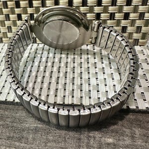 May include: A silver-toned wristwatch with an expandable band. The watch face is not visible, but the back casing is round and metallic. The band is made of small, rectangular metal links that stretch to fit the wrist. The watch is set against a textured background.