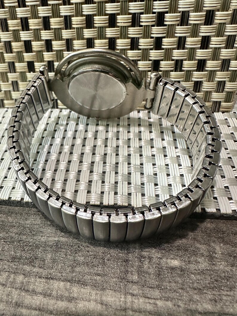 May include: A silver-toned wristwatch with an expandable band. The watch face is not visible, but the back casing is round and metallic. The band is made of small, rectangular metal links that stretch to fit the wrist. The watch is set against a textured background.