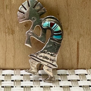 Puede incluir: Una figura de Kokopelli plateada, un símbolo nativo americano, con incrustaciones de piedras turquesas y rojas. La figura toca una flauta, con diseños grabados detallados en la ropa y el tocado. El fondo es una superficie de madera clara.