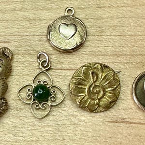 May include: A collection of vintage gold-toned jewelry pieces, including a bear-shaped brooch, a heart-shaped locket, a pendant with a green stone, a flower-shaped brooch with a musical note, and a round brooch with turquoise and white accents.