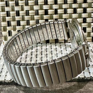 May include: A silver-toned, stainless steel watch with an expandable band. The band is made of rectangular metal links. The watch face is visible, and the band is curved. The watch is on a textured surface.