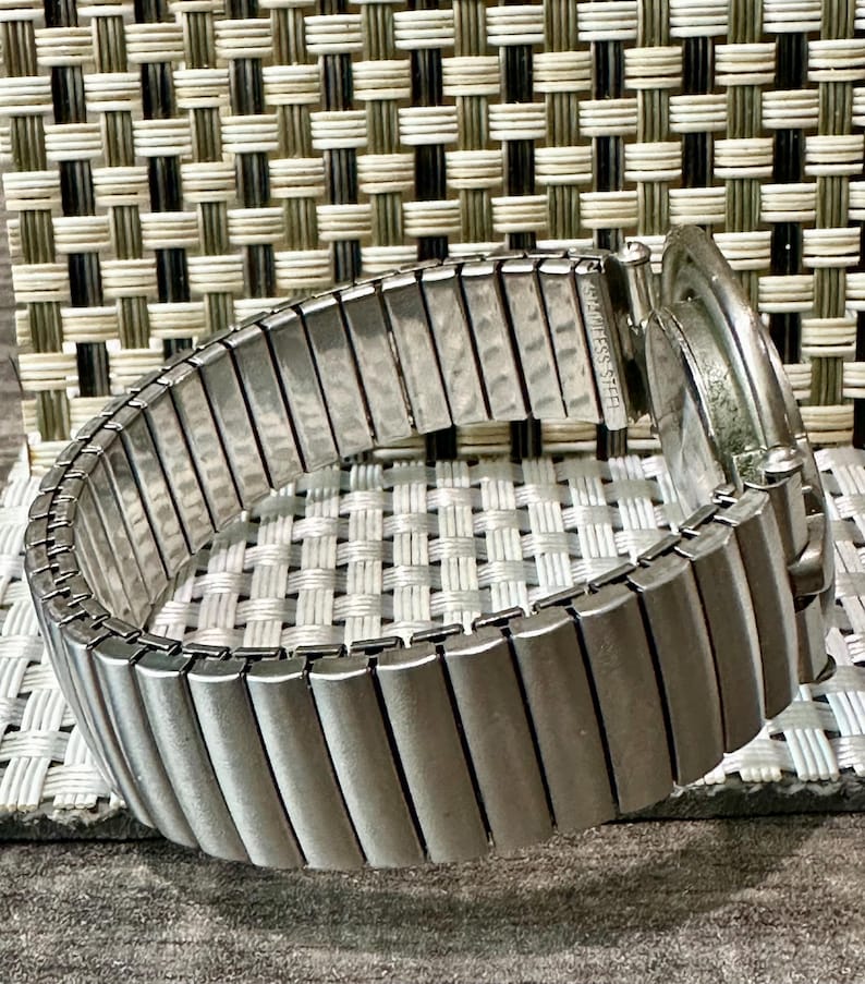 May include: A silver-toned, stainless steel watch with an expandable band. The band is made of rectangular metal links. The watch face is visible, and the band is curved. The watch is on a textured surface.