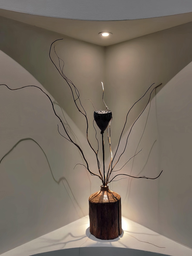 May include: A dried lotus pod in a wooden vase with thin branches extending from the vase. The vase and branches are in a corner of a room with a white ceiling and white walls.
