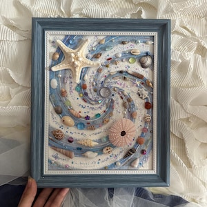 Könnte beinhalten: Gerahmtes Kunstwerk mit maritimem Thema, das ein spiralförmiges Design aus Muscheln, Seesternen und bunten Perlen auf einem weiß-blauen Hintergrund zeigt. Der Rahmen ist in einem verwitterten Blau gehalten. Das Kunstwerk umfasst verschiedene Muscheltypen und -größen.