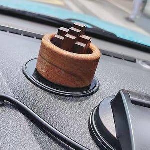 Puede incluir: Un soporte de teléfono para coche de madera con una base cilíndrica y un interior marrón oscuro, que contiene varios bloques de madera rectangulares pequeños. El soporte se encuentra sobre una base circular negra, montado en el salpicadero del coche.