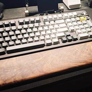 Puede incluir: Un teclado mecánico con reposamuñecas de madera. El teclado tiene teclas grises y blancas. El reposamuñecas es de madera con un patrón de vetas natural. Un ratón negro es visible a la derecha del teclado.