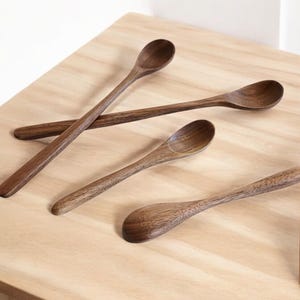 Könnte beinhalten: Vier Holzlöffel mit dunkelbraunem Finish sind auf einer hellen Holzoberfläche angeordnet. Die Löffel variieren in Größe und Form, mit länglichen Griffen und abgerundeten Schalen. Die Löffel bestehen aus Naturholz.