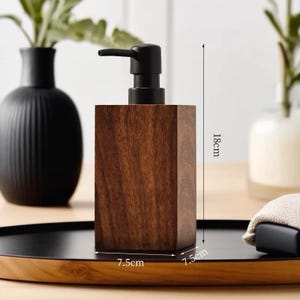 Puede incluir: Un dispensador de jabón de madera marrón oscuro con una bomba negra. El dispensador tiene 7,5 cm de ancho y 7,5 cm de profundidad. Tiene 18 cm de alto.