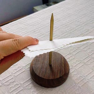 Puede incluir: Una base de madera con una varilla de metal dorado y una hoja de papel blanca unida a la varilla. La hoja de papel está siendo sostenida por una mano.
