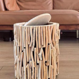 Puede incluir: Una mesa auxiliar cilíndrica de madera con tapa. La mesa está construida con numerosos palitos de madera pequeños y de color claro, creando una estética texturizada y natural. La tapa es de madera clara similar. Mueble decorativo.