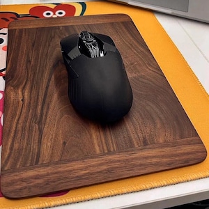 Op de afbeelding: Een zwarte gamingmuis met een textuur zit op een donkerbruine houten muismat met een lichtbruine rand. De muismat ligt op een geel stoffen oppervlak.