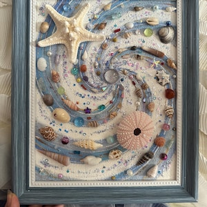 Puede incluir: Obra de arte enmarcada con un diseño en espiral, que incorpora arena, conchas marinas y elementos decorativos. Una gran estrella de mar está en la parte superior, con varias conchas, cuentas y un erizo de mar rosa. El marco es azul claro.
