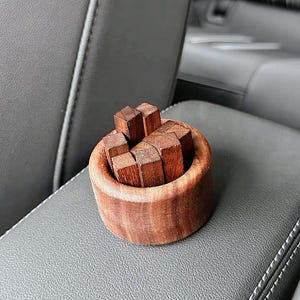 May include: A small, round wooden container filled with several small, square wooden blocks. The container and blocks are a rich, reddish-brown color. The items are sitting on a gray surface.