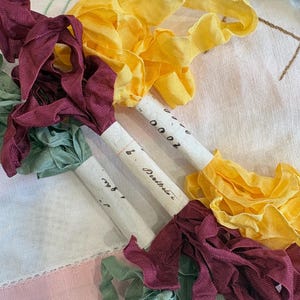 May include: Three bundles of fabric ribbon in yellow, deep burgundy, and sage green. Each bundle is secured with a white paper band featuring handwritten text. The ribbons have a textured, woven appearance and are arranged in a loose, overlapping cluster.