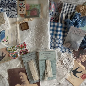 May include: A collection of vintage ephemera and textiles including blue and white patterned fabrics, embroidered linens, lace trims labelled Au Bon Marche and Au Printemps, antique postcards, a small bag of buttons, and blue silk ribbons.