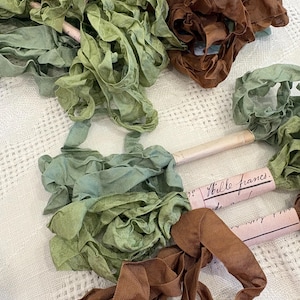 May include: A selection of ruffled ribbons in shades of green and brown. Some ribbons are rolled on small cardboard tubes with handwritten text. The ribbons are arranged on a textured, off-white surface.