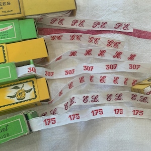 May include: A collection of vintage ribbon spools and printed ribbons. The ribbons are white with red text and numbers. The spools are in green and yellow boxes with French text.