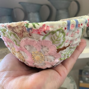 Pode incluir: Uma tigela pequena, feita à mão, feita de sobras de tecido. A tigela é decorada com bordados florais rosa, branco e verde.