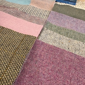 May include: A collection of rectangular fabric swatches featuring various textures and patterns, including solid pink, olive green, purple, and herringbone weaves. The swatches are arranged in a grid-like layout alongside a stack of five blue books.