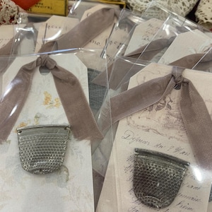 May include: Two antique silver thimbles with a floral design on the tag. The tag is tied with a brown ribbon and has the text "Dejeuner du 7 Janv. 1877" written on it.