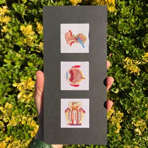 May include: A black card featuring three cross-stitch squares depicting anatomical diagrams of the ear, eye, and tooth. The cross-stitch designs use red, blue, yellow, and brown threads on a white background. The card is held against a backdrop of green and yellow foliage.