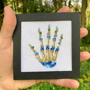 Op de afbeelding: Een kruissteekkunstwerk met een skelet handontwerp. De hand is gestikt met bruin, blauw en groen garen op witte stof, omlijst in een zwart vierkant. Het kunstwerk wordt in een hand gehouden tegen een vage groene achtergrond.