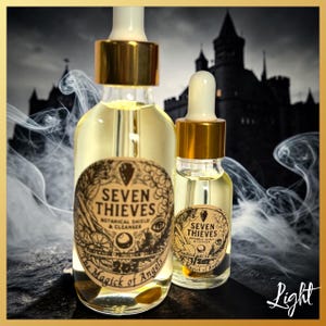 May include: Two clear glass dropper bottles with gold caps containing a light yellow liquid. Each bottle features a round beige label with black text reading Seven Thieves Botanical Shield & Cleanser The Magick of Angels.