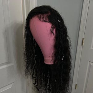 May include: A black curly human hair wig on a pink mannequin head. The wig is styled in loose curls and has a lace front.