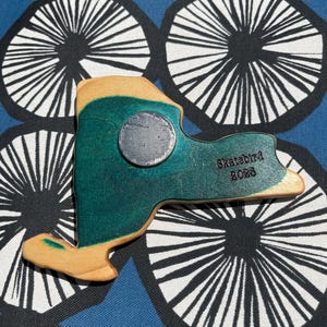 May include: A wooden magnet in the shape of New York State, painted teal and tan. A silver magnet is attached to the front. The text "Skatebird 2023" is printed on the back. The magnet is on a blue patterned surface.