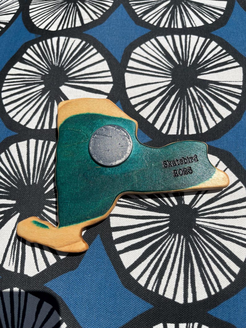 May include: A wooden magnet in the shape of New York State, painted teal and tan. A silver magnet is attached to the front. The text "Skatebird 2023" is printed on the back. The magnet is on a blue patterned surface.
