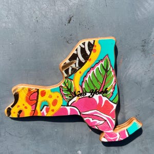 May include: A wooden New York state-shaped decorative item with a vibrant floral design. The artwork features a pink rose, green leaves, and yellow and orange patterns, with the words "New York" in cursive.