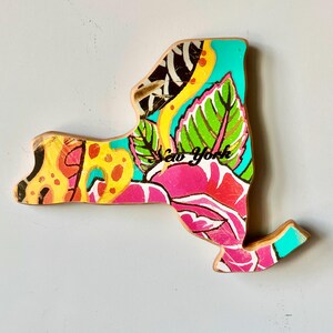 May include: Wooden New York state cutout with a vibrant floral design. The artwork features a mix of colors, including pink, yellow, green, and teal. The words "New York" are written in black script across the design.