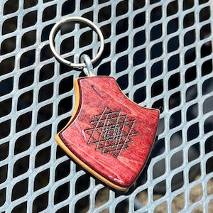 May include: A red wooden keychain with a geometric design etched into the surface. The keychain has a silver metal ring and a light-colored wood border. The geometric design is a series of interlocking triangles.