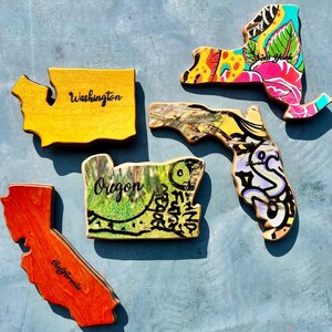 May include: Wooden state-shaped art pieces. The states of Washington, Oregon, California, New York, and Florida are depicted. Each piece is painted with unique designs and colors, including floral patterns and graffiti-style art. The word "Washington", "Oregon", "California", and "New York" are written on the pieces.