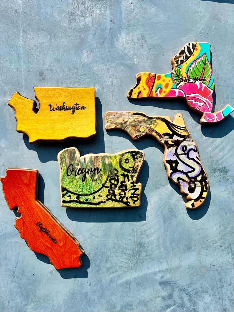 May include: Wooden state-shaped art pieces. The states of Washington, Oregon, California, New York, and Florida are depicted. Each piece is painted with unique designs and colors, including floral patterns and graffiti-style art. The word "Washington", "Oregon", "California", and "New York" are written on the pieces.