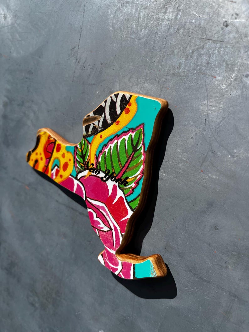 May include: Wooden New York state-shaped magnet with a colorful floral design. The magnet features a vibrant mix of colors, including pink, teal, yellow, and green, with the words "New York" in cursive. The edges are a natural wood color.