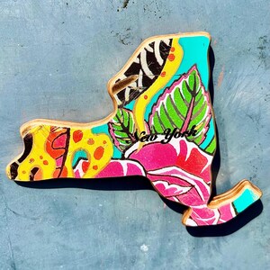 May include: Wooden New York state cutout with a vibrant floral design. The artwork features a mix of colors, including yellow, pink, green, and turquoise, with the words "New York" in pink script. The cutout is on a blue surface.