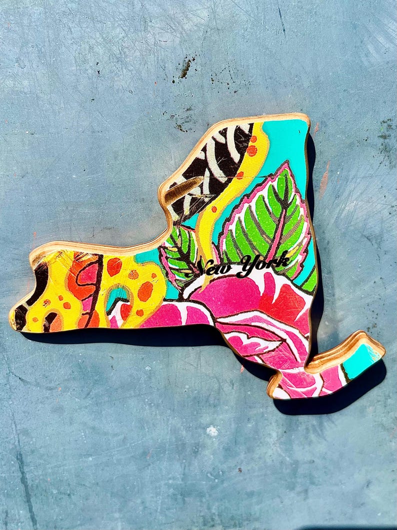 May include: Wooden New York state cutout with a vibrant floral design. The artwork features a mix of colors, including yellow, pink, green, and turquoise, with the words "New York" in pink script. The cutout is on a blue surface.