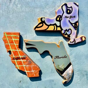 May include: Three wooden state-shaped art pieces: New York, California, and Florida. The New York piece is purple with black and yellow accents and the text "New York". California is orange with a green grid and the text "California". Florida is gray with the text "Florida".