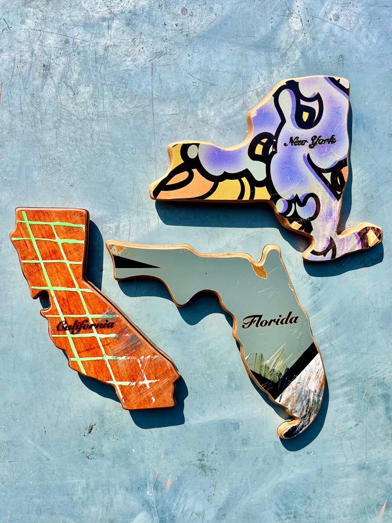 May include: Three wooden state-shaped art pieces: New York, California, and Florida. The New York piece is purple with black and yellow accents and the text "New York". California is orange with a green grid and the text "California". Florida is gray with the text "Florida".