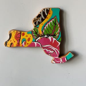 May include: Wooden New York state cutout with a colorful floral design. The artwork features a vibrant mix of pink, yellow, green, and teal, with the words "New York" written across the center.
