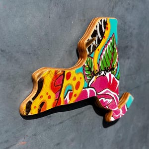 May include: Wooden New York state-shaped magnet with a colorful floral design. The magnet features a vibrant mix of yellow, pink, blue, and green, with the text "New York" incorporated into the artwork.