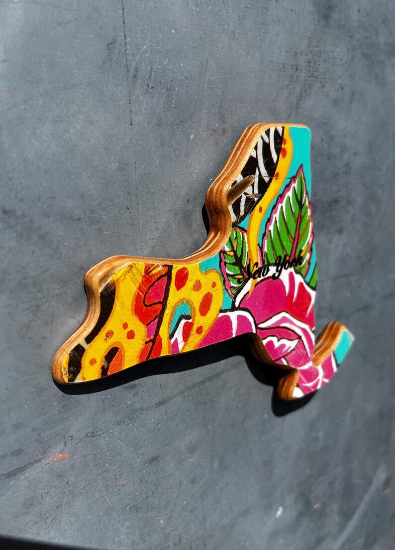 May include: Wooden New York state-shaped magnet with a colorful floral design. The magnet features a vibrant mix of yellow, pink, blue, and green, with the text "New York" incorporated into the artwork.