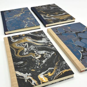 May include: Four notebooks with marbled covers in shades of blue, black, gold, and white. The notebooks have a variety of cover designs, with some featuring a blue marbled pattern and others a black and gold design. The spines are a natural tan color.