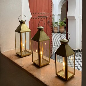 May include: Three brass lanterns with glass panels and lit candles are on a windowsill. The lanterns have a classic design with a handle on top. The background features a red door and a courtyard with black and white tiles.