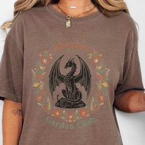 以下が含まれることがあります： エスプレッソ色のTシャツにドラゴンのグラフィック。デザインは、黒いドラゴンが花のリースに囲まれ、「Lotus Garden Club」の文字が入っています。 ゆったりとしたフィット感のシャツです。