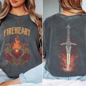 Op de afbeelding: Donkergrijs t-shirt met het woord "FIREHEART" in gouden letters. Het ontwerp aan de voorkant toont een hart, vlammen en rozen. De achterkant van het shirt toont een zwaard omhuld door vlammen.