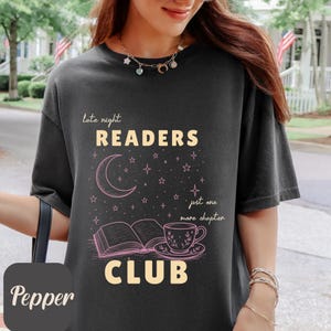 Könnte beinhalten: Ein anthrazitfarbenes T-Shirt mit einem Grafikdruck, der den Text "late night READERS CLUB" und "just one more chapter" zeigt. Das Design umfasst eine Mondsichel, Sterne, ein offenes Buch und eine Teetasse mit Untertasse in hellem Rosa.