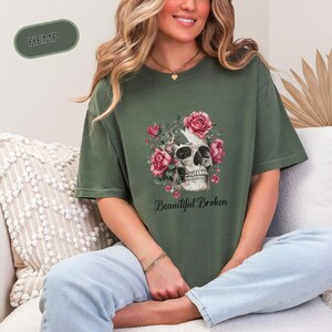 Könnte beinhalten: Olivgrünes T-Shirt mit einem Totenkopf-Design, das mit rosa Rosen verziert ist. Die Worte "Beautiful Broken" sind unter dem Totenkopf aufgedruckt. Das Shirt hat eine lockere Passform und ist aus weichem Material.