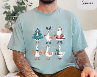 May include: Dusty blue t-shirt with a festive design featuring geese dressed as Christmas characters. The geese are depicted as a Christmas tree, reindeer, Santa Claus, and with gingerbread and gifts. The shirt is labeled "Bella Canvas".
